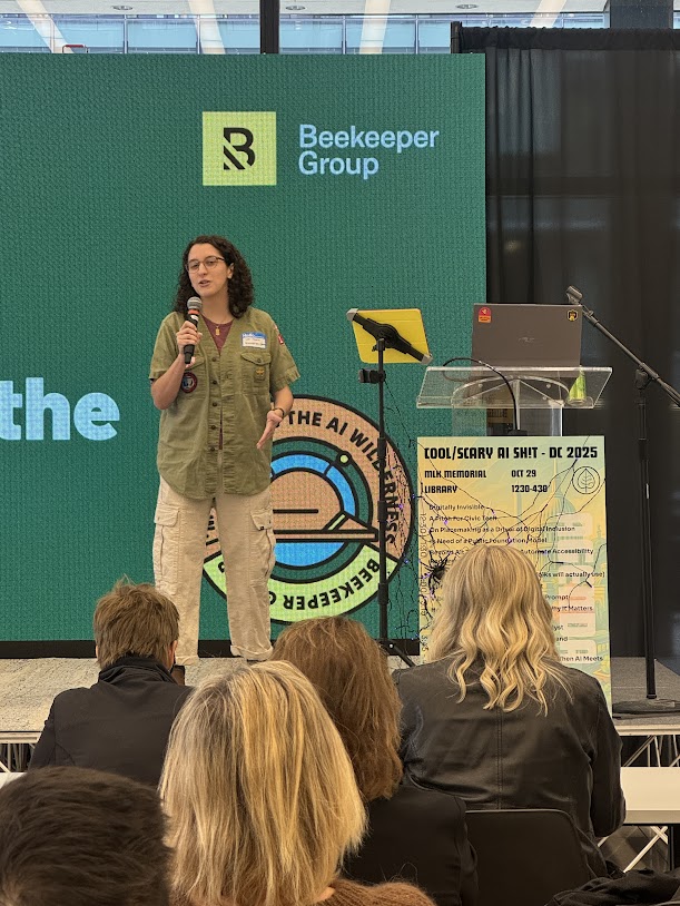 A person stands on stage holding a microphone, recapping Beekeeper Group's 2025 day of purpose. Behind them, a large screen displays the Beekeeper Group logo and a colorful event poster beside a table with a laptop.