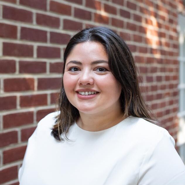A woman with long brown hair wearing a white top stands in front of a red brick wall, smiling at the camera. Sunlight creates patches of light behind her—an inviting scene that captures the welcoming spirit of Beekeeper Group.
