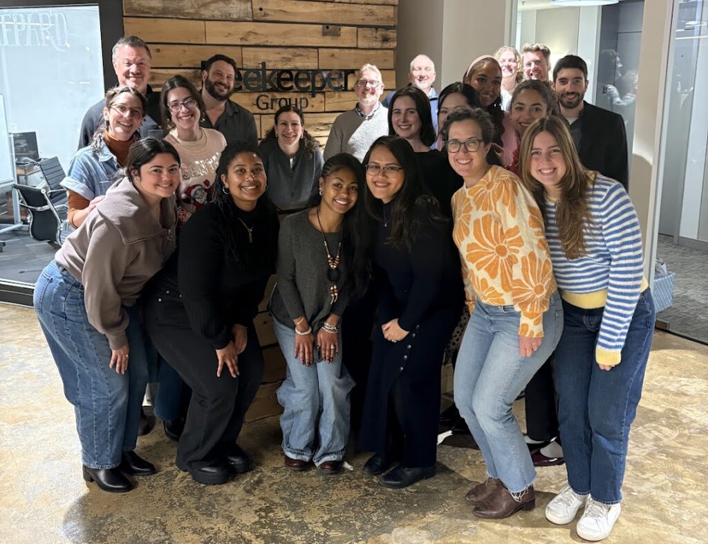 A group of about 20 people smiling and posing together in an office space with wooden paneling and glass walls. The team stands closely in front of a wall with the sign about Beekeeper Group visible.