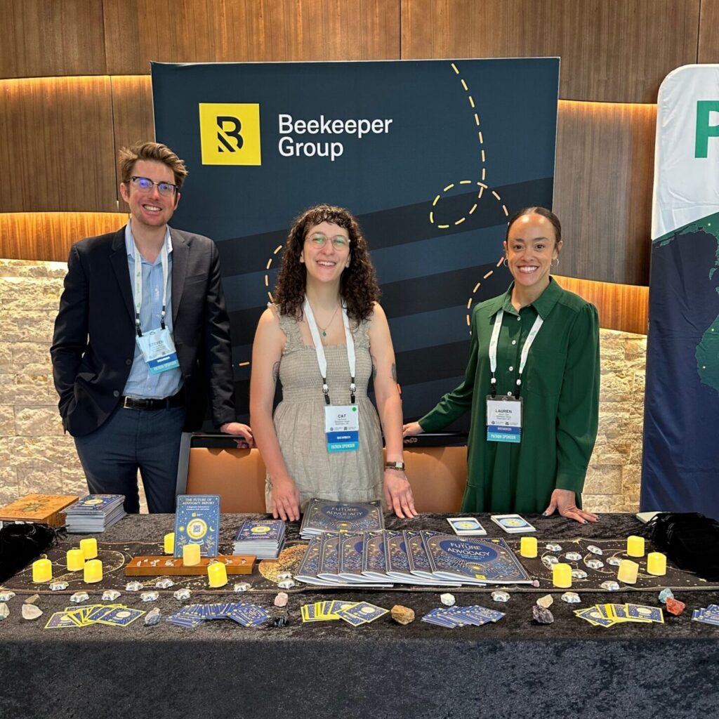 Three people stand smiling behind a table covered with cards, candles, and promotional items at a Beekeeper Group booth, featuring an annual recap from Beekeeper Group on display beneath a yellow and black banner against a wood-paneled wall.