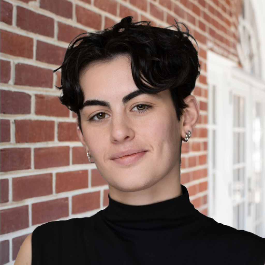 A person with short dark hair, wearing a black sleeveless turtleneck and hoop earrings, smiles slightly while standing in front of a red brick wall and white windowed doors—perfect for the Spring 2026 Hive Five Newsletter.