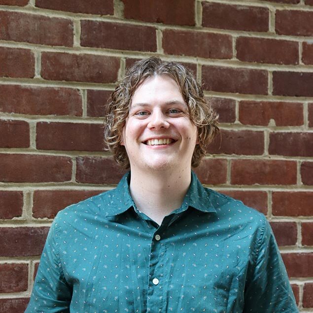 Sam Keimweiss, with curly light brown hair, is smiling while standing in front of a red brick wall, wearing a green short-sleeved button-up shirt with a subtle pattern.