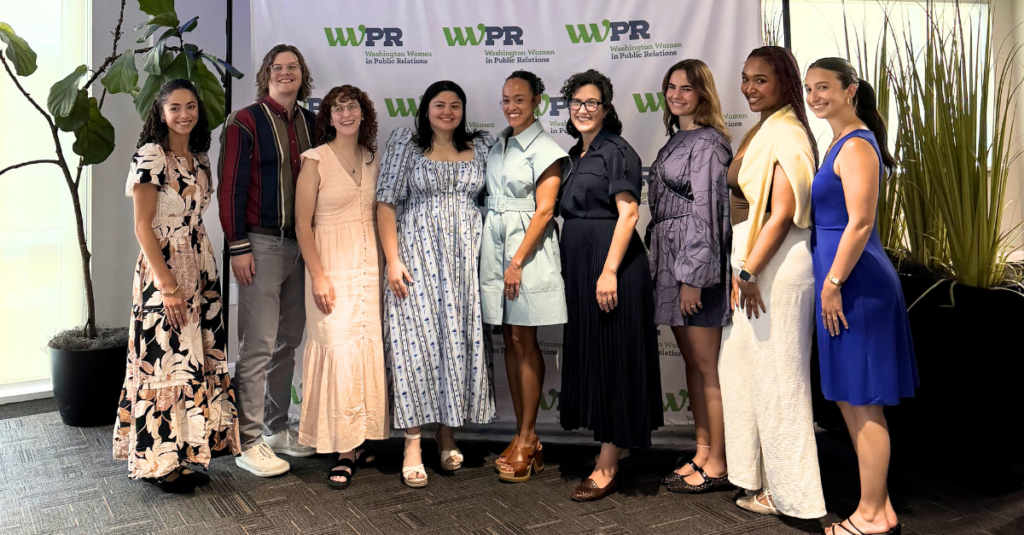 Ten people stand smiling in front of a WNPR/WPR backdrop, dressed in business casual and semi-formal attire. Some wear dresses, others suits or coordinated outfits, perfectly embodying the energy of Summer 2026. Large plants decorate the left and right sides of the photo, capturing a moment featured in the Hive Five Newsletter.