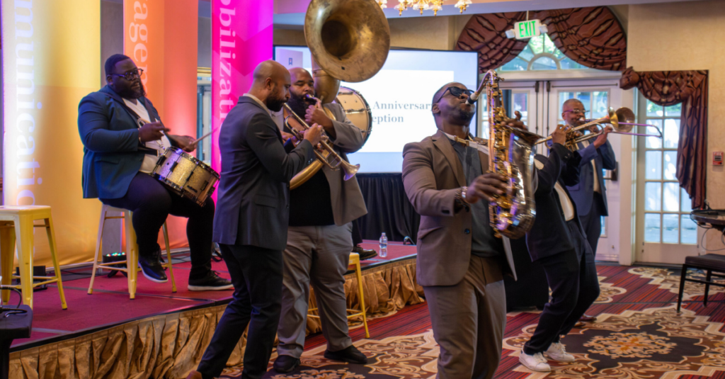A jazz band plays instruments on stage, including drums, trumpet, trombone, and saxophone, in front of a colorful backdrop and audience at an indoor event—perfect for featuring in the Summer 2026 edition of the Hive Five Newsletter.