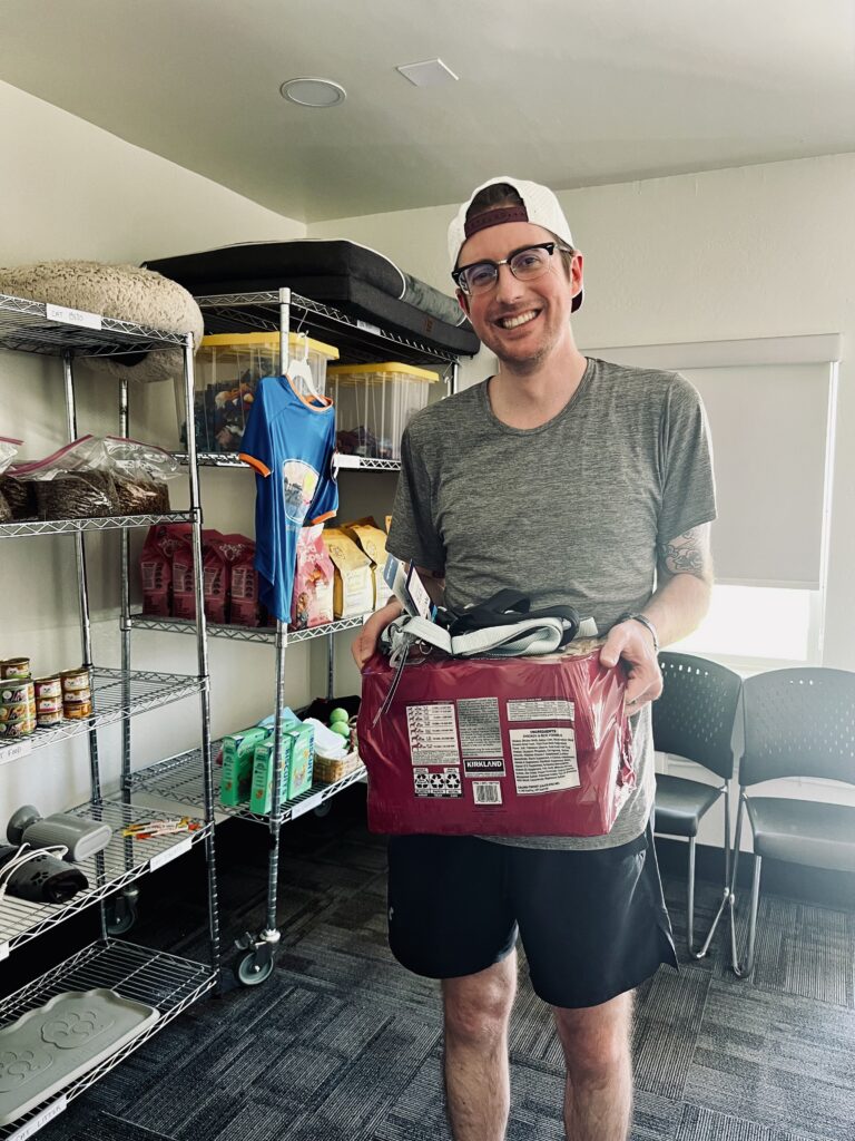 A smiling person wearing glasses, a gray shirt, and black shorts holds a bag of pet food in a room with shelves of various supplies and a row of chairs, recapping Beekeeper Group’s 2025 day of purpose.