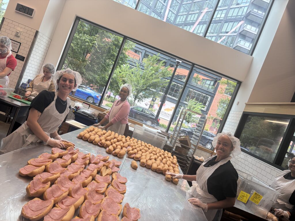 People wearing aprons and hairnets prepare sandwiches in a bright kitchen with large windows and city views. Rows of sandwiches are on metal tables, and everyone is smiling while working together, recapping Beekeeper Group’s 2025 day of purpose.
