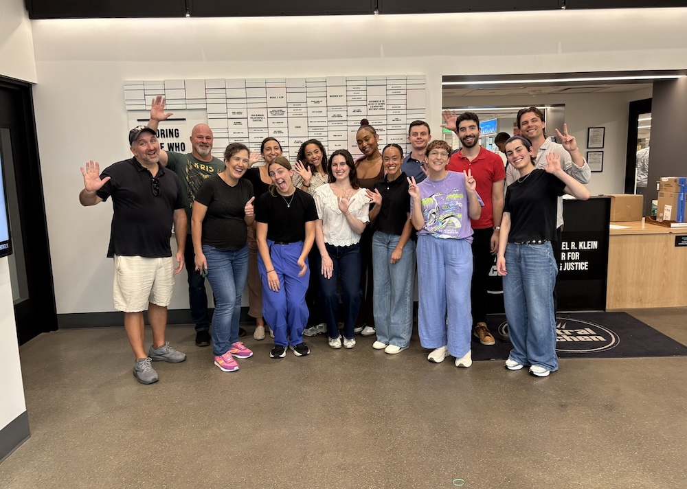A group of fifteen people pose together indoors, smiling and waving at the camera. They are casually dressed and standing in front of a wall with a large white schedule board, recapping Beekeeper Group’s 2025 day of purpose.